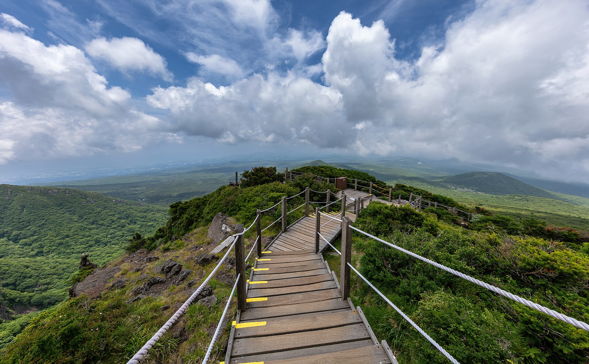 Wooden staircase along Yeongsil Trail showing a panoramic view on the green subtropical vegetation and the mountains of Hallasan National Park , UNESCO Biosphere Reserve and World Heritage Site of Jeju Island , in South Korea .