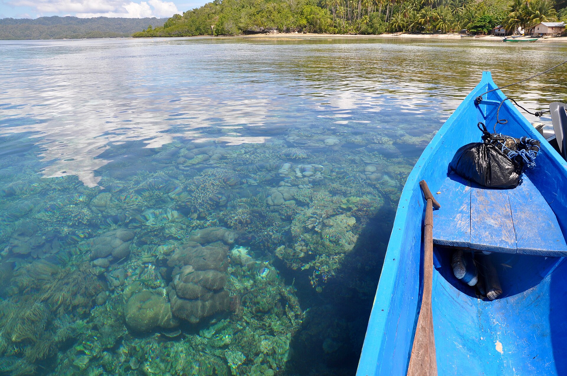 A narrow local boat floating above clear water and shallow coral near Gam Island in Raja Ampat